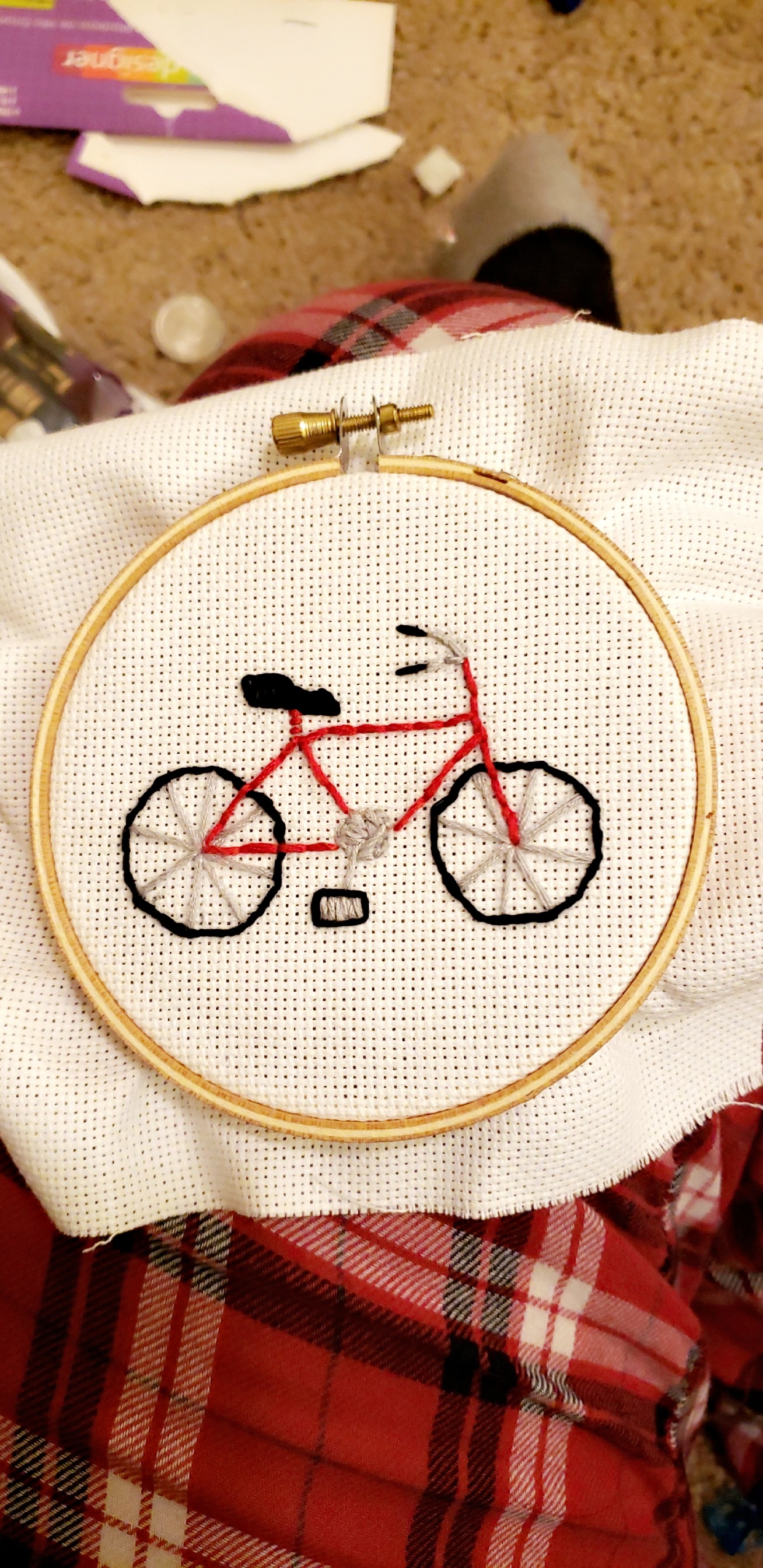 an image of white aida cloth with a red bicycle stitched in embroidery floss in a circular embroidery hoopresting on someones leg